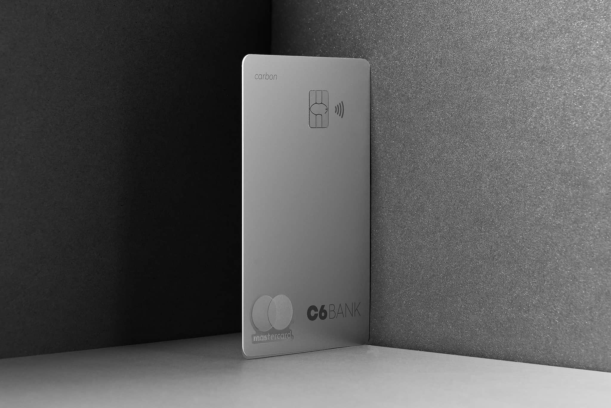 Foto do cartão C6 Carbon Mastercard Black prateado na vertical apoiado em uma cartolina em tons de cinza