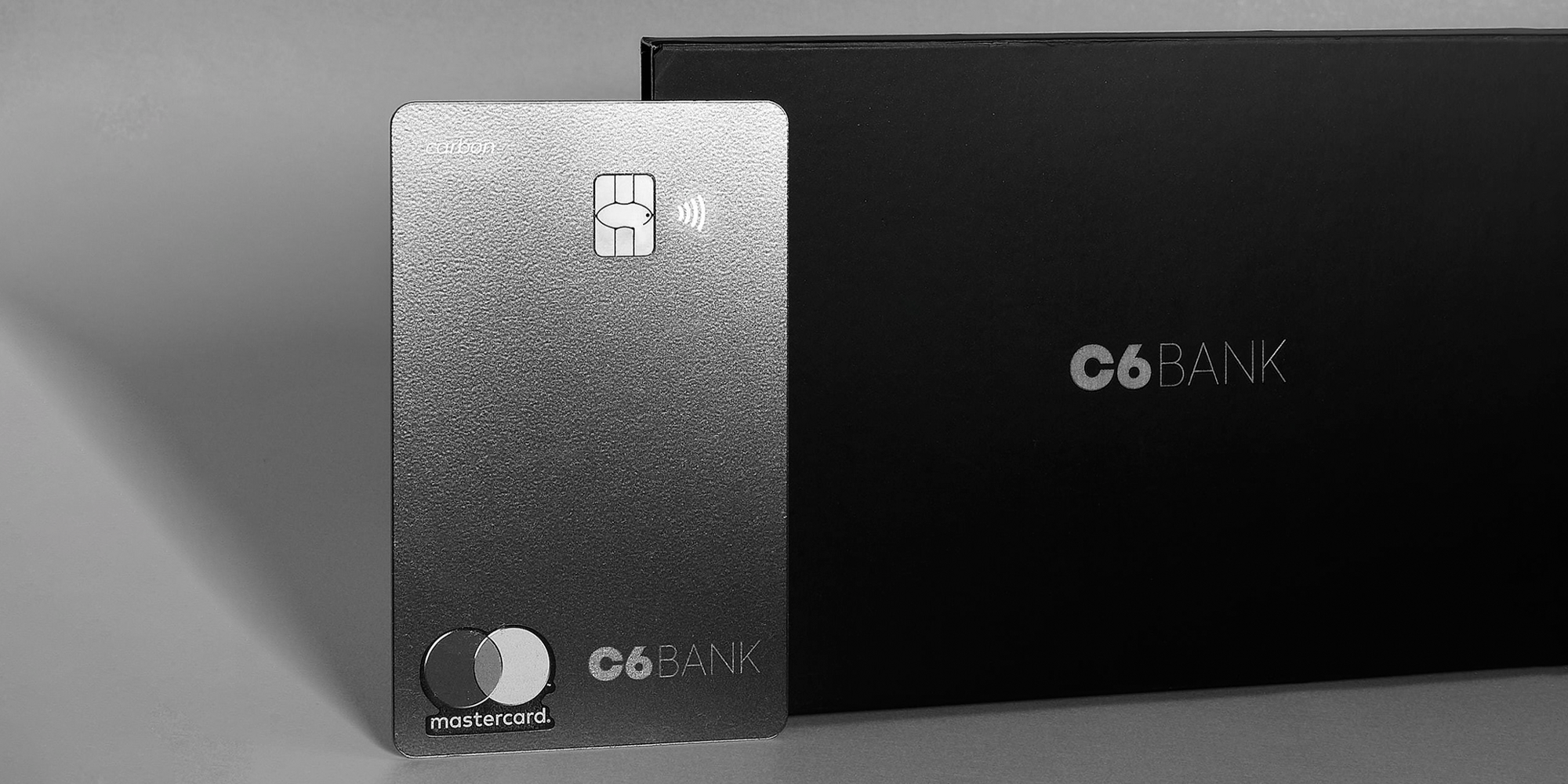Foto do cartão C6 Carbon Mastercard Black na vertical apoiado no envelope em que ele é enviado para a casa dos clientes