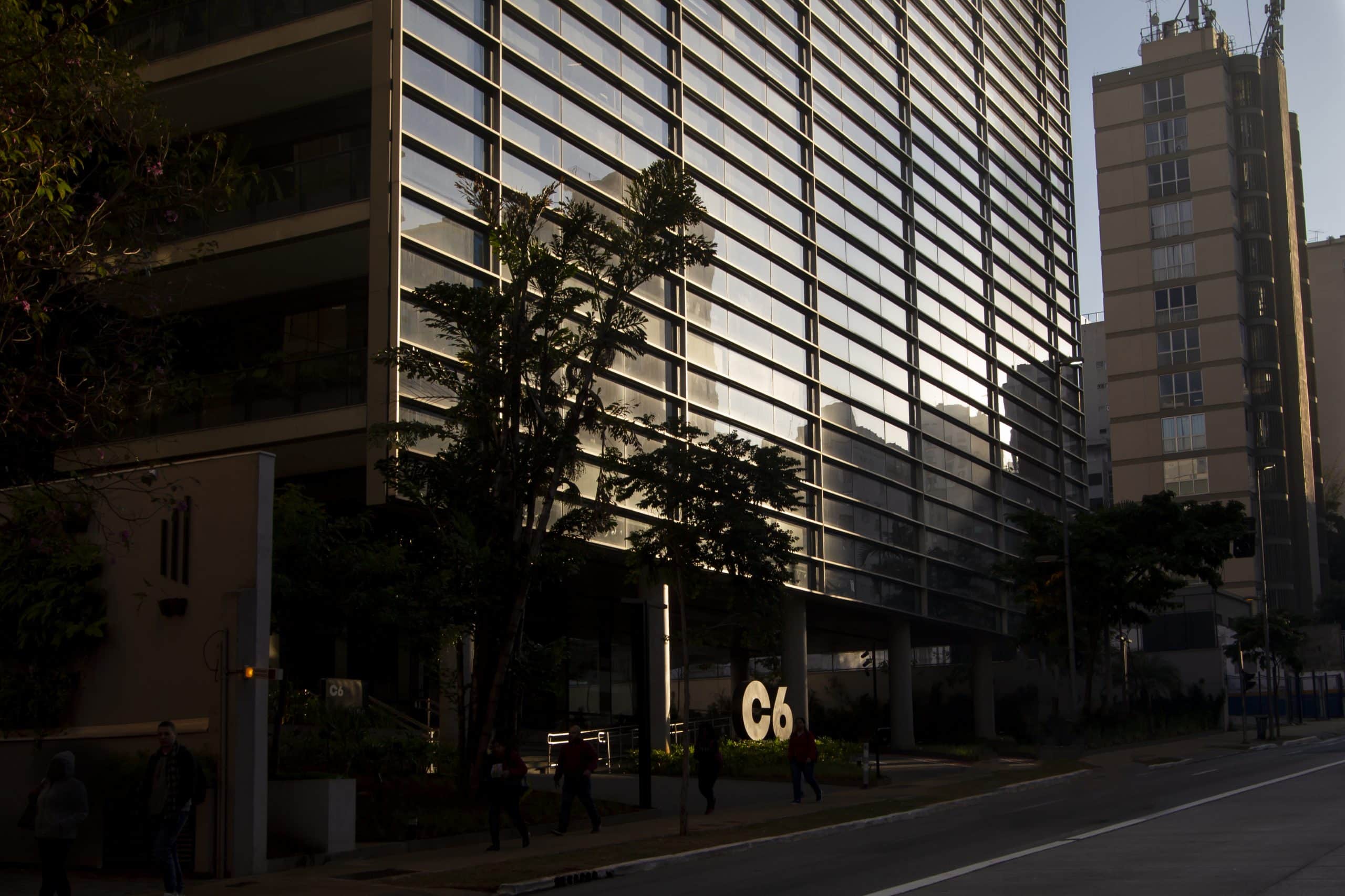 Foto da fachada do prédio do C6 Bank onde se pode ver o logo do banco brilhando com a luz do sol, enquanto algumas pessoas caminham pela calçada