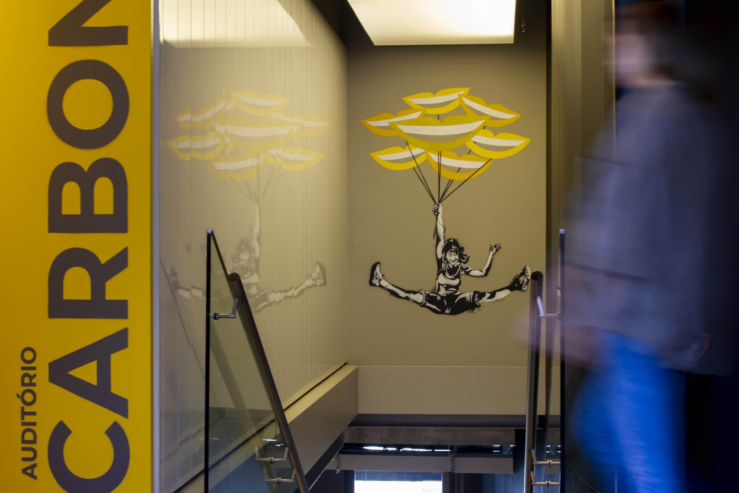 Foto de um grafismo na parede do prédio do C6 Bank de uma garota sorrindo e voando ao segurar balões em formato de sorriso na cor amarela