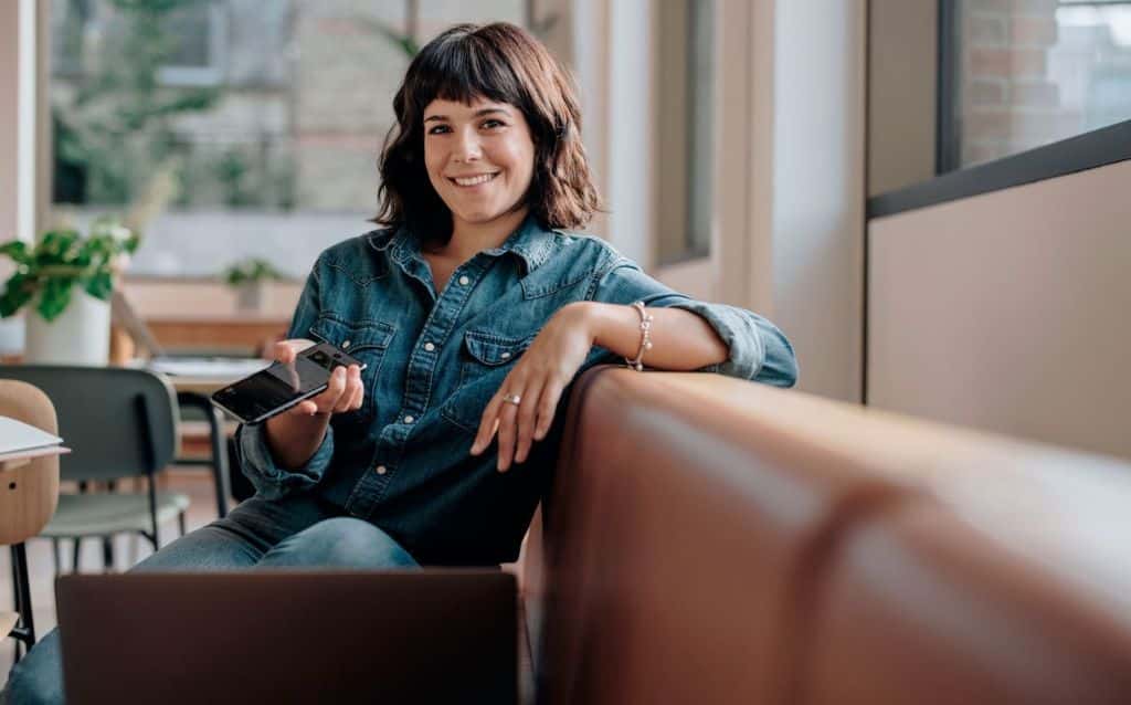 mulher com celular aberto em carteira digital na mão, está sentada em um sofá de couro marrom em um estabelecimento comercial