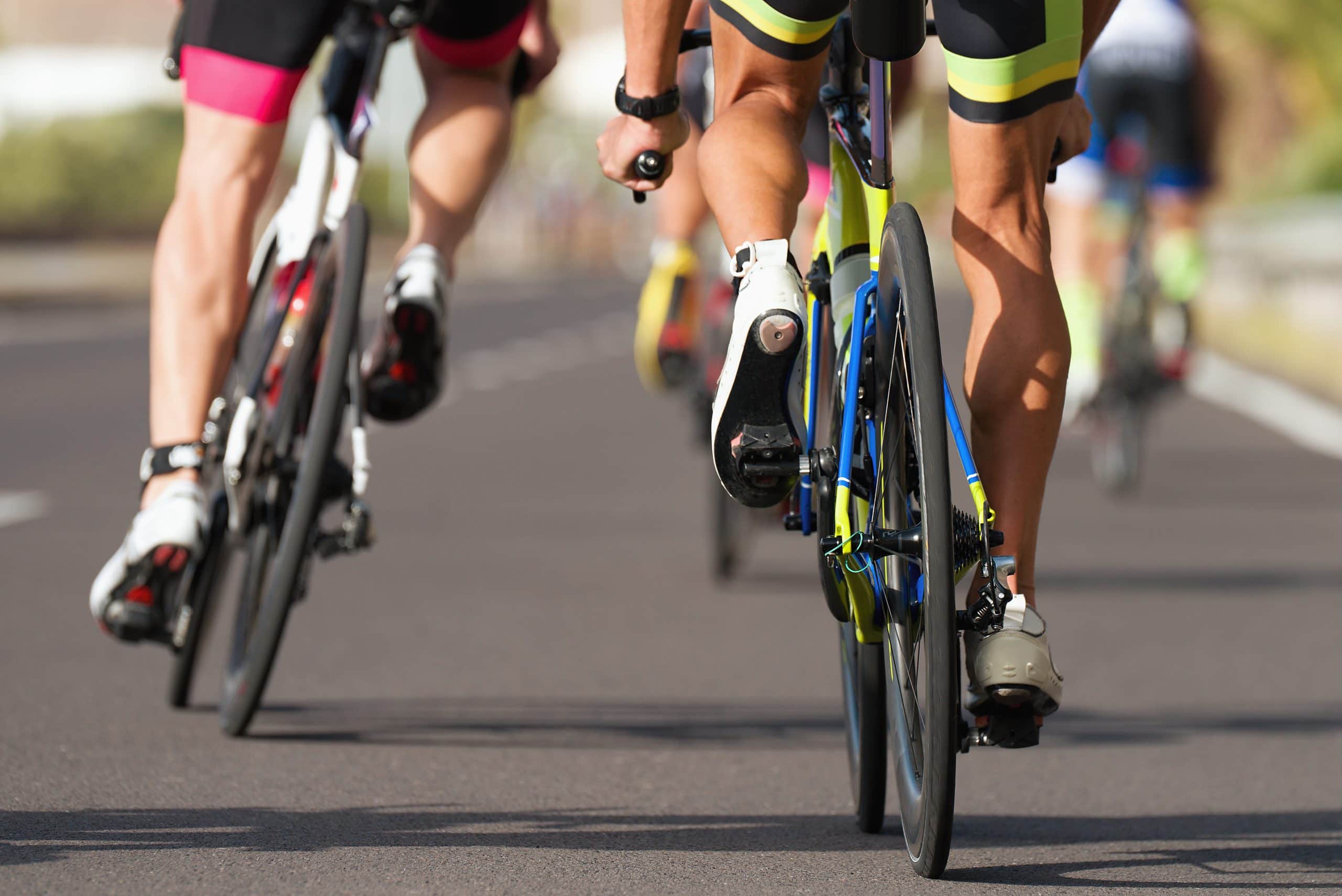 Foto de dois ciclistas pedalando em bicicletas e trajes de corrida. Os ciclistas estão de costas. As bicicletas estão no asfalto. Em primeiro plano, o ciclista usa sapatilha branca e uma bermuda preta com faixa amarela. Em segundo plano, um ciclista usa sapatilha branca, com bermuda preta e faixa rosa.
