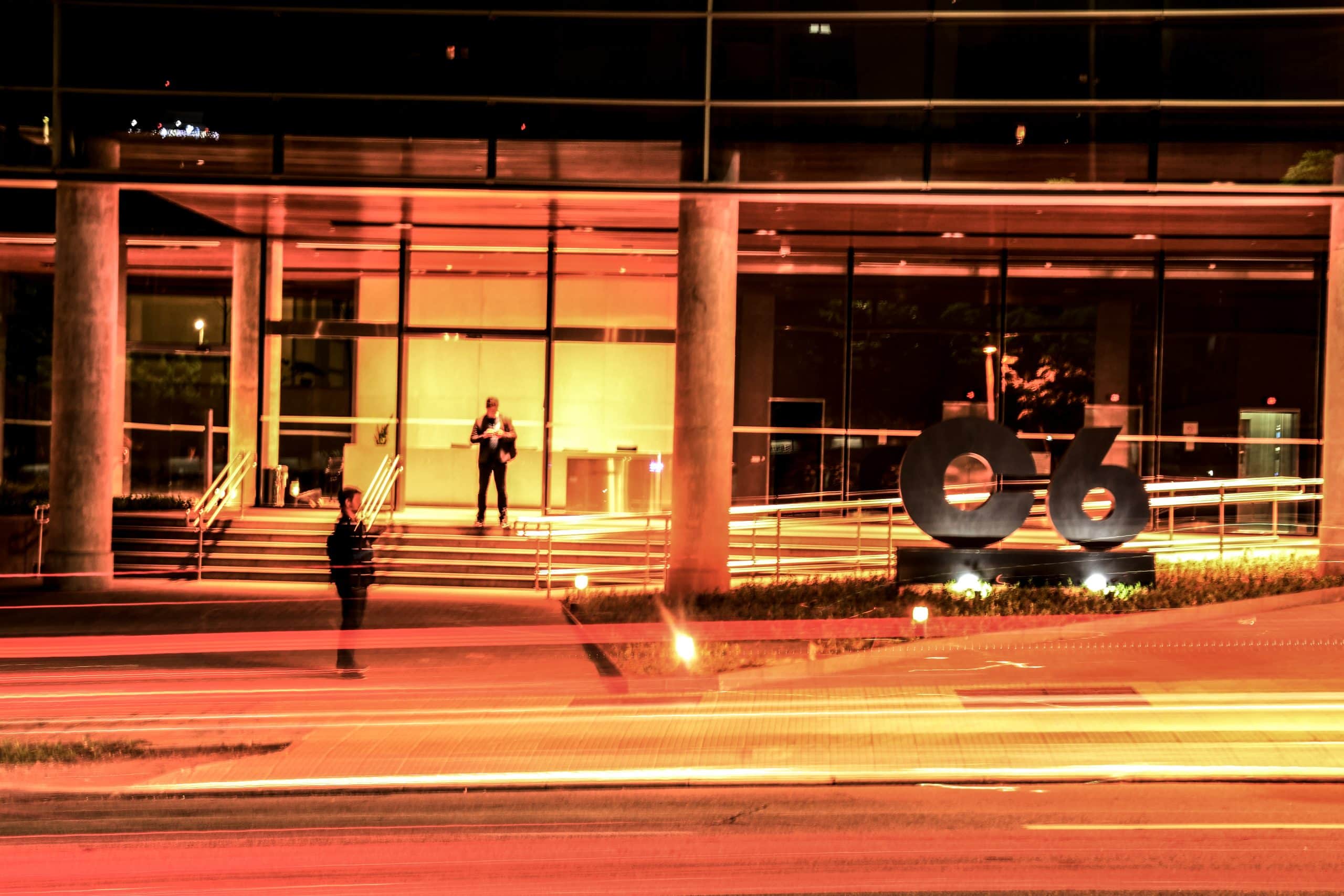 Foto da fachada do prédio do C6 Bank durante a noite com time lapse de carros passando na avenida em frente com luzes amarelas
