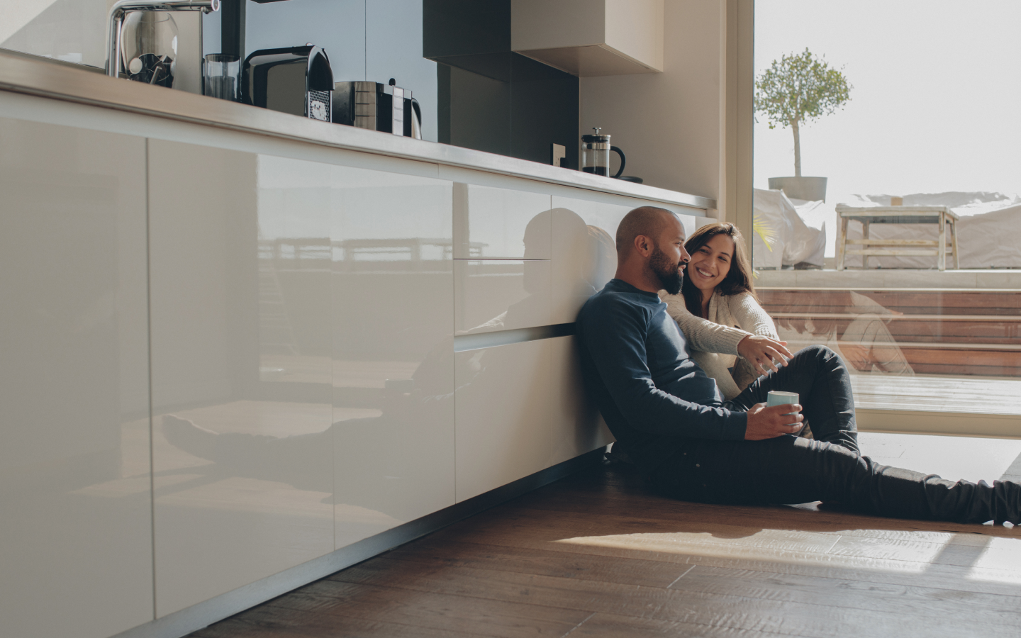 casal, formado por um homem e uma mulher brancos, sentados no chão de uma cozinha conversando, de forma a simbolizar o planejamento financeiro familiar