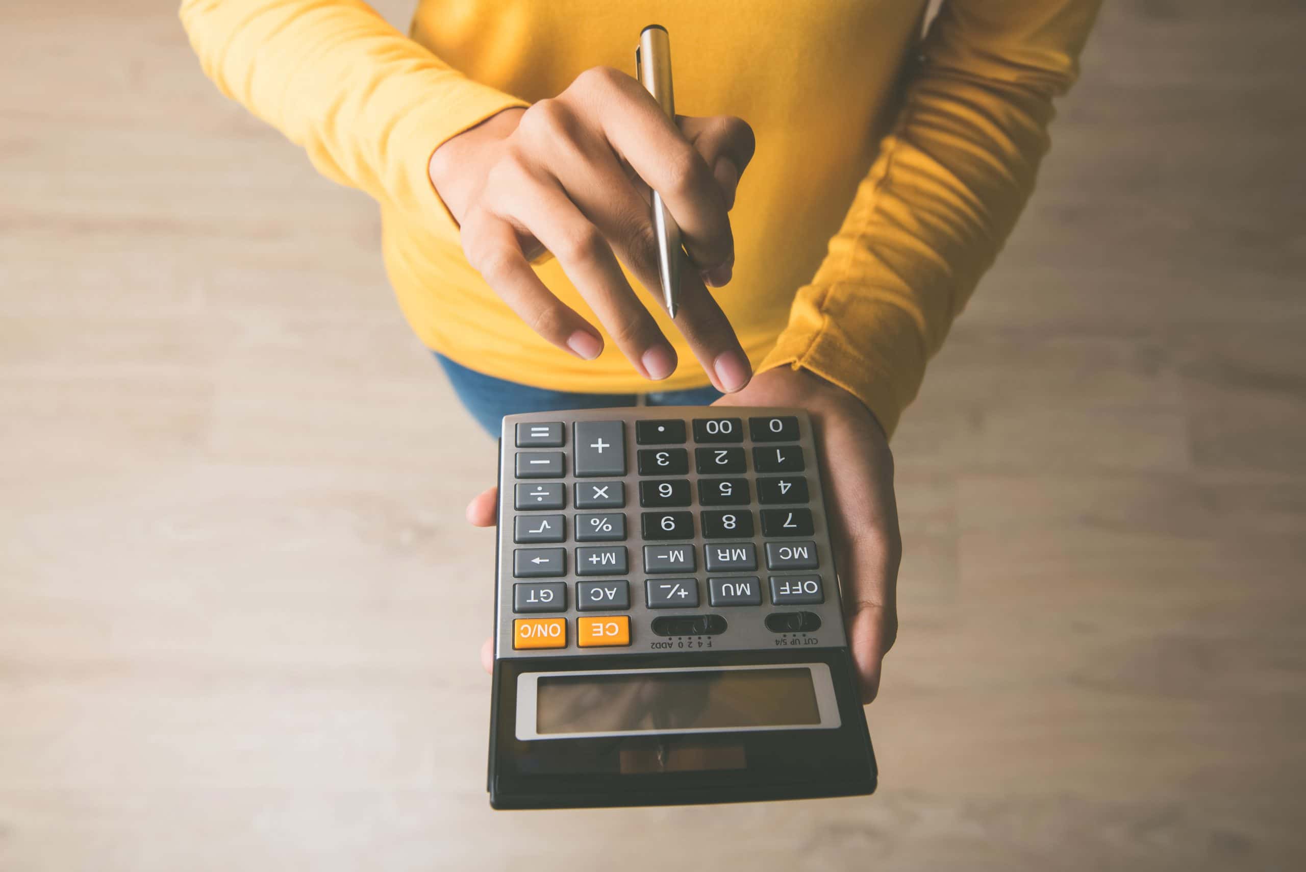 Pessoa de blusa de manga longa amarela, segurando uma calculadora em uma mão e uma caneta na outra
